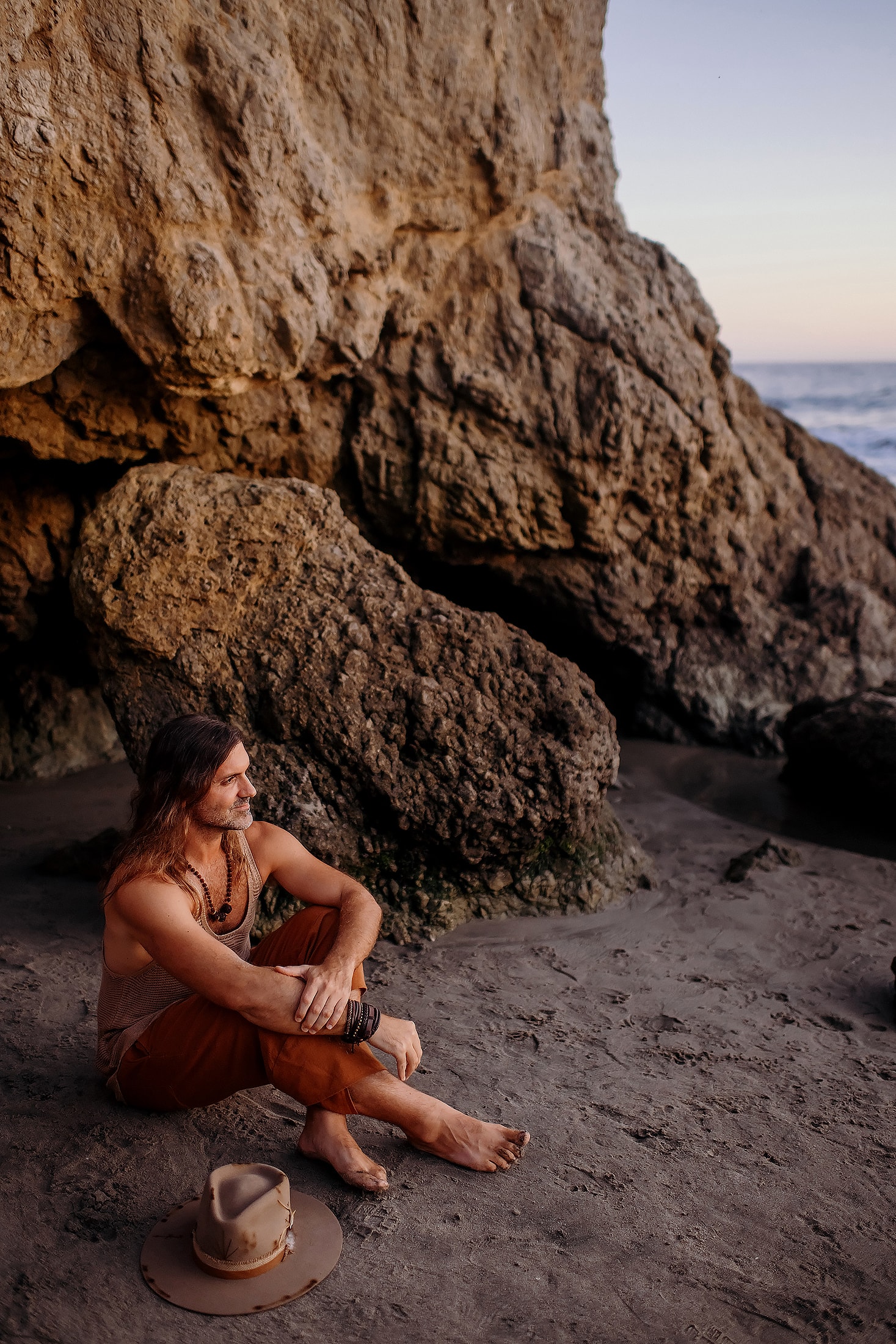 Daniel Bellone sitting on the beach at golden hour beside the ocean, his hat resting on the sand